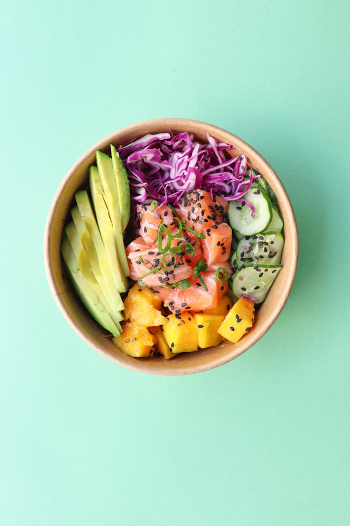 about-img Top view of healthy poke bowl with fresh blue cabbage with chopped avocado cucumber and mango with raw salmon on green background
