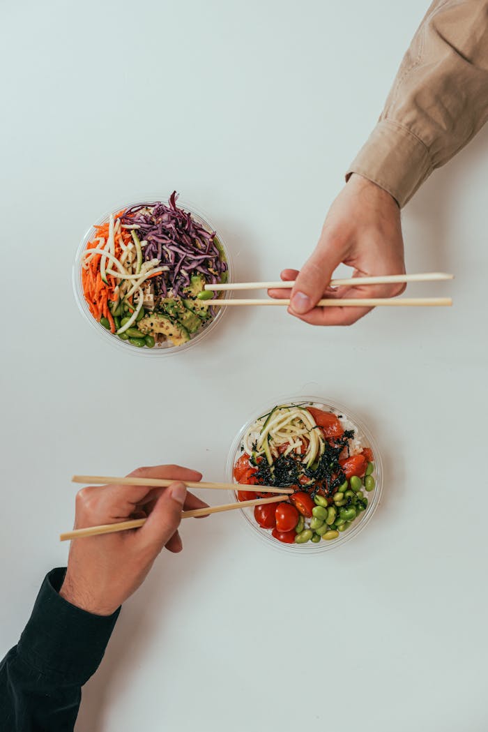 gallery-2 Two people using chopsticks to enjoy fresh vegetable bowls on a white table.