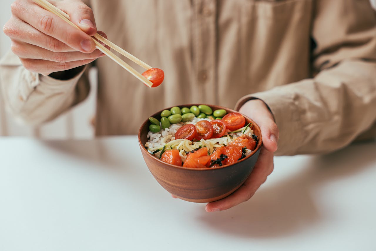 gallery-6 A person holding a rice bowl with cherry tomatoes, edamame, and more, using chopsticks.